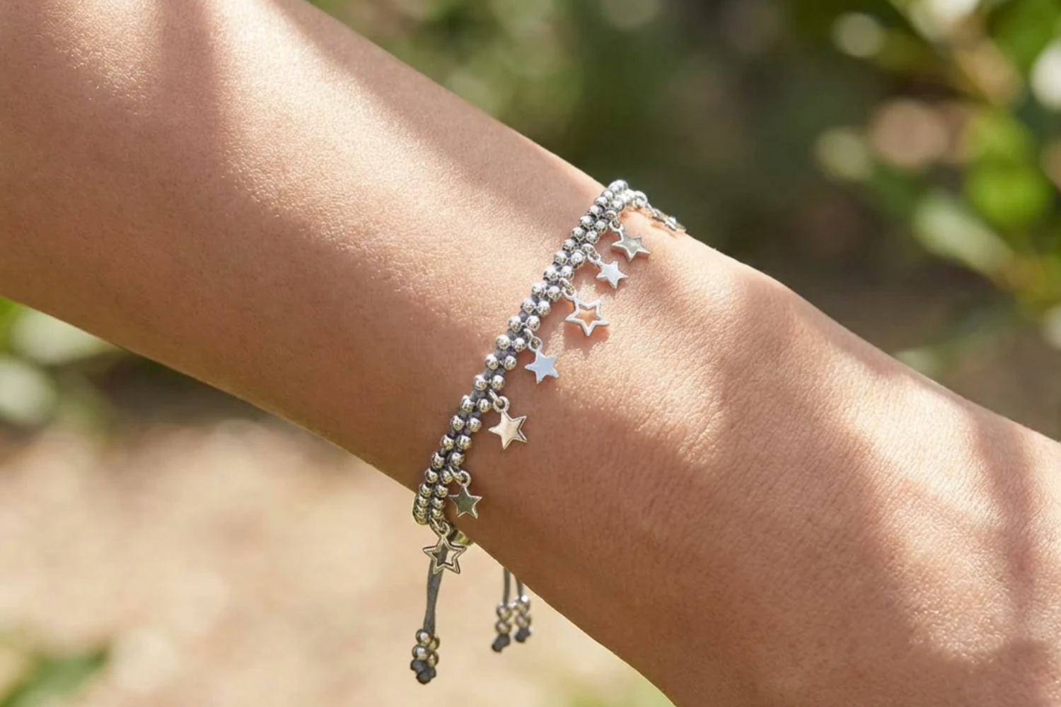 Close-up of a female model's wrist showcasing the Estelle Grey & Silver Star Charm Bracelet with grey cord, silver beads, and star charms in natural outdoor lighting.