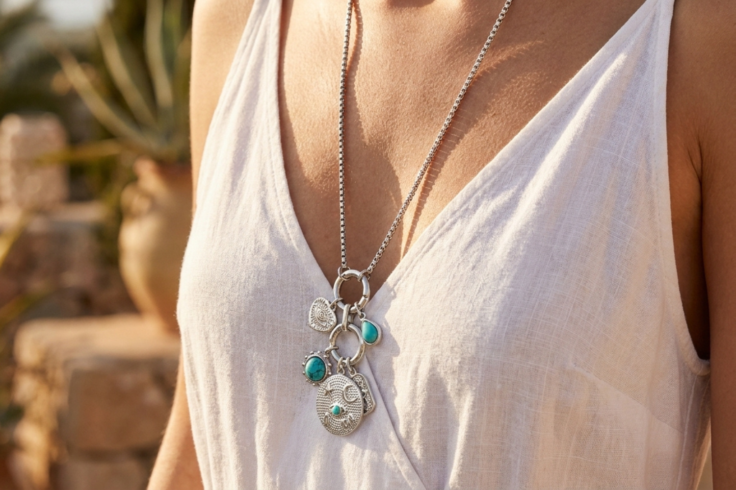 Close-up of a female model wearing a long silver necklace with detachable charms including turquoise howlite stones, over a summer dress, showcasing the detailed chain and charms in a breezy outdoor setting.