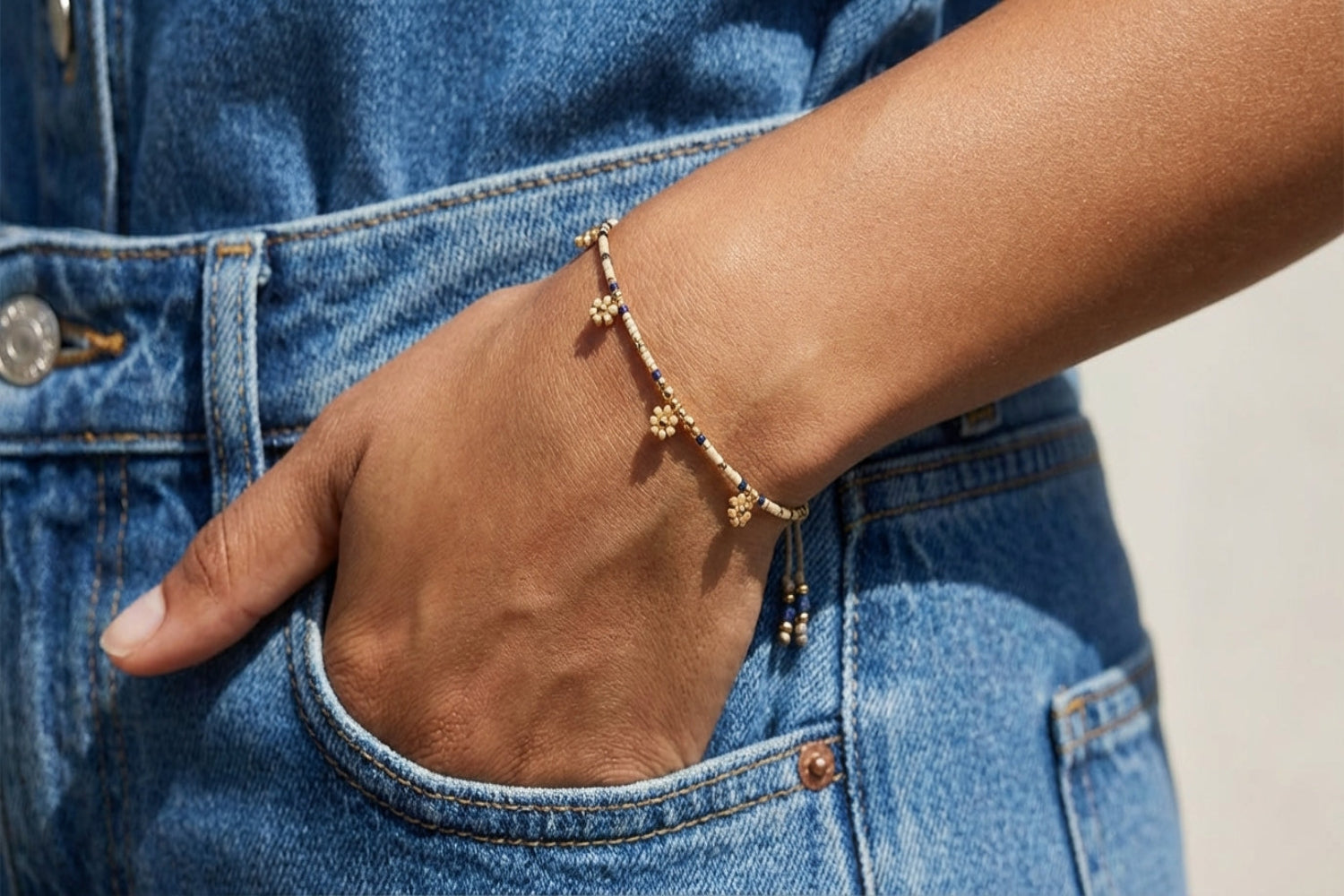 Close-up of a female model with medium skin tone wearing the Rania Blue & Gold Beaded Charm Bracelet on her arm, with her hand in a denim pocket, showing detailed beadwork and floral charms.