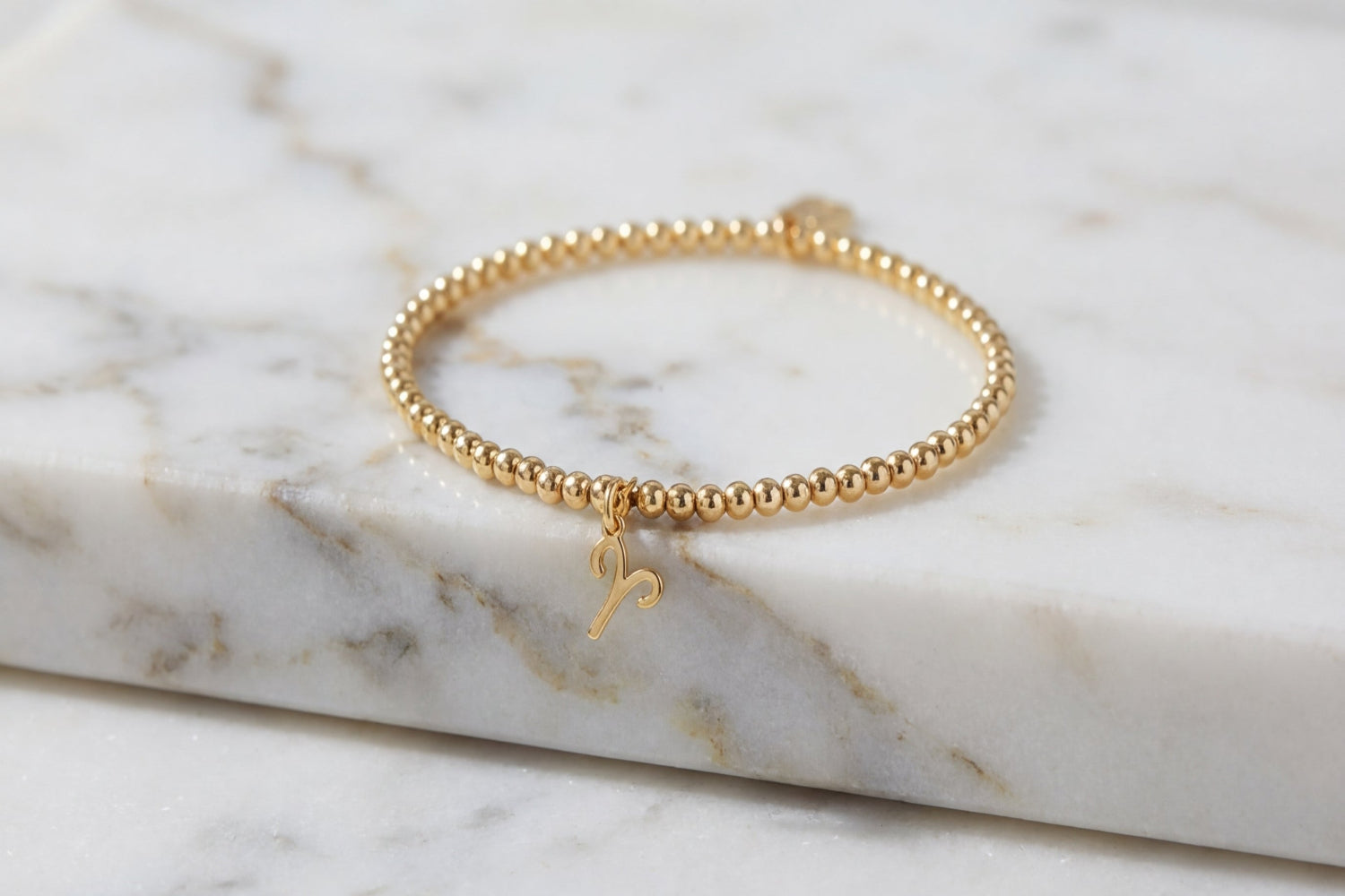 Close-up of a delicate gold Aries zodiac charm bead bracelet on a marble surface, highlighting the intricate Aries symbol and fine stretch beads.