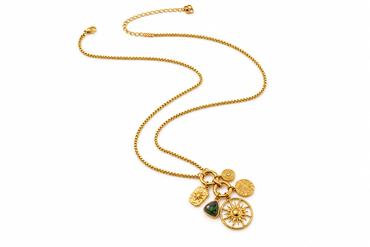 A gold chain necklace with multiple small dangling charms, including a sunburst wheel, a green stone, and two round coin-like discs, shown on a plain white background.