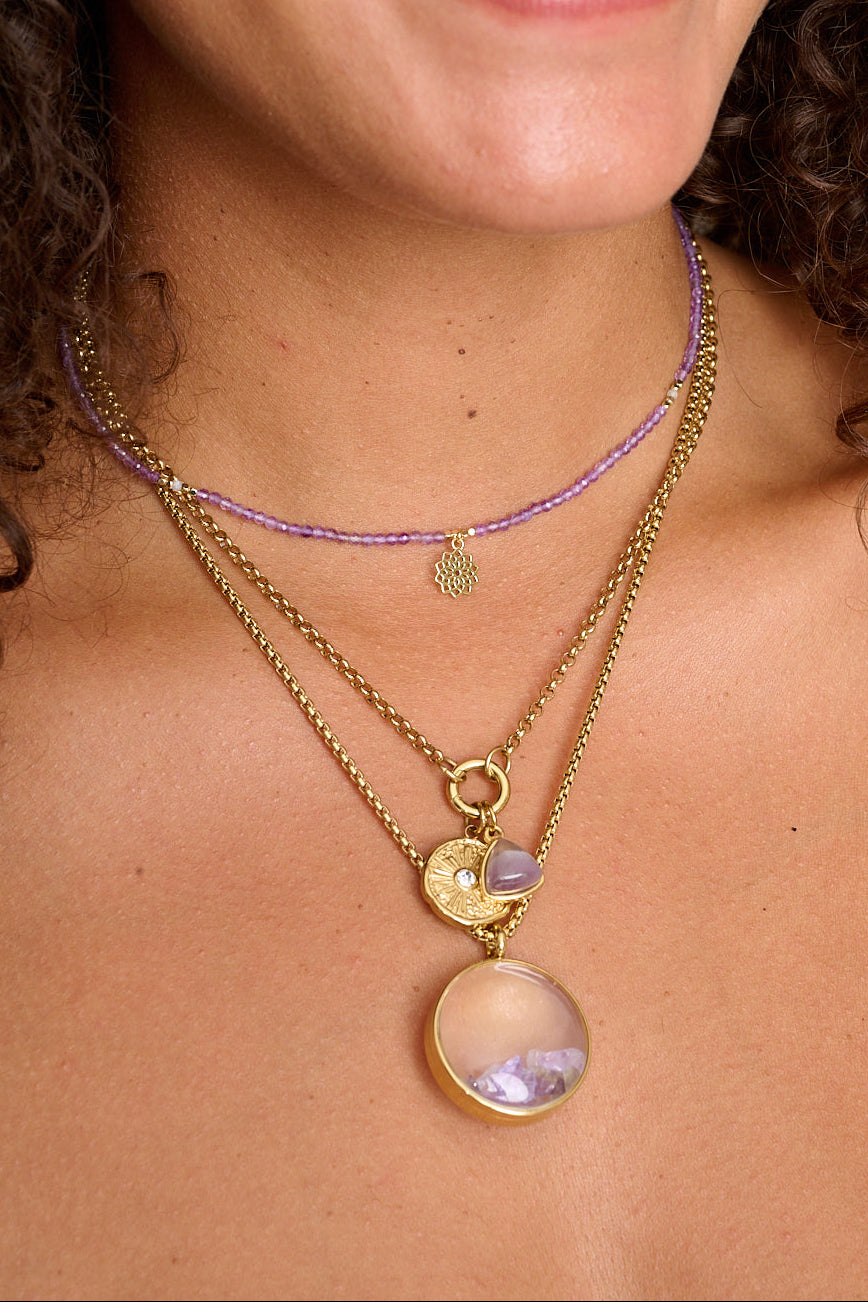 Leilani Gold Locket Necklace styled with a layered gold chain stack, showcasing gemstone chips and letter charms inside the round glass pendant.