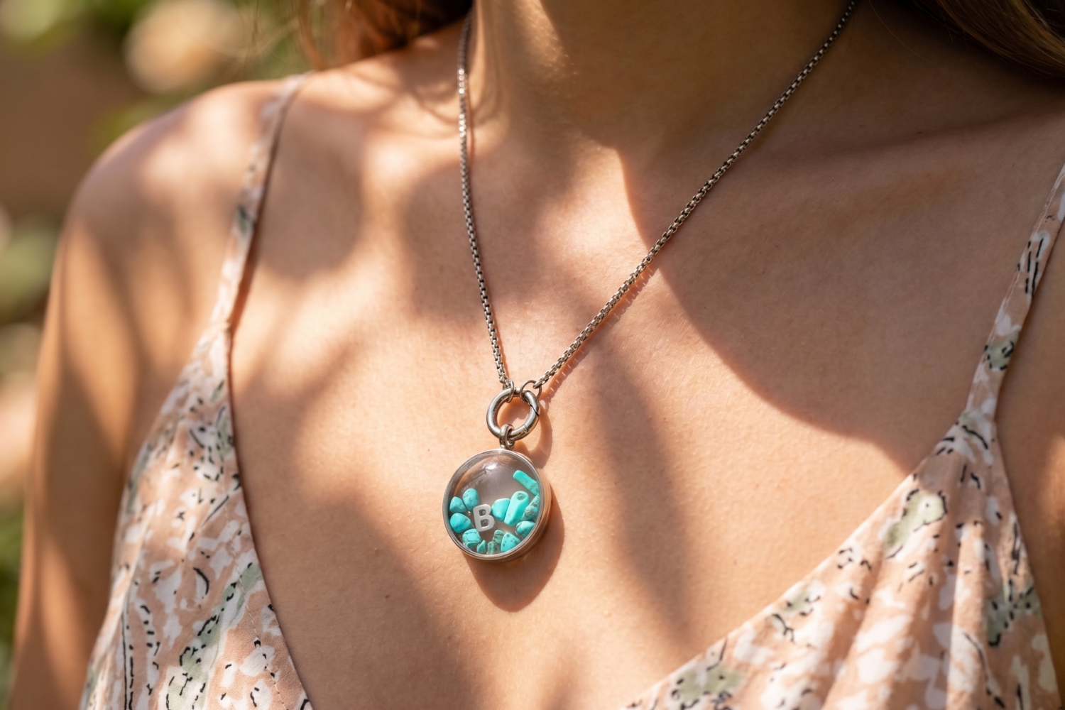 Close-up of a female model wearing a stainless steel letter locket necklace with gemstone chips, styled longer for summer fashion, face-on shot.