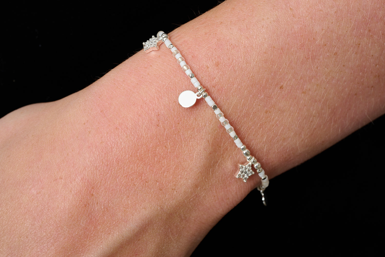 Stack of bracelets featuring the Rania White & Silver Beaded Star Charm Bracelet paired with silver accents and soft-toned outfits.