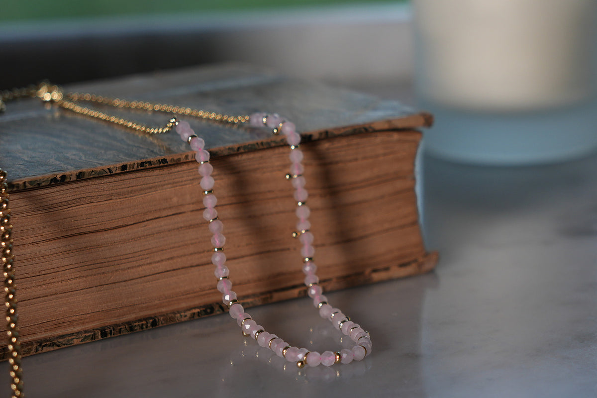 Salus Rose Quartz Gemstone Gold Necklace of faceted pale pink rose quartz beads and gold accents draped over an antique book