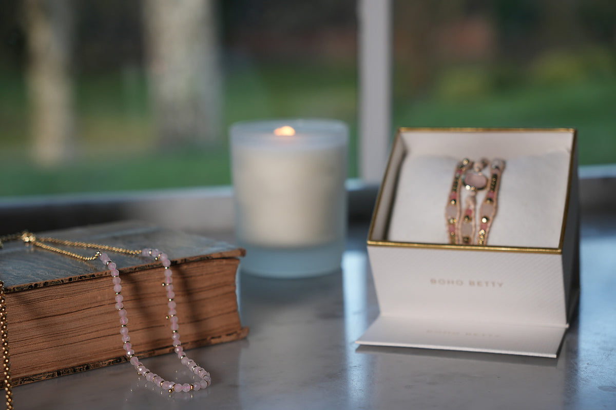 Salus Rose Quartz Gemstone Gold Necklace draped over a vintage book beside a candle and a Boho Betty gift box