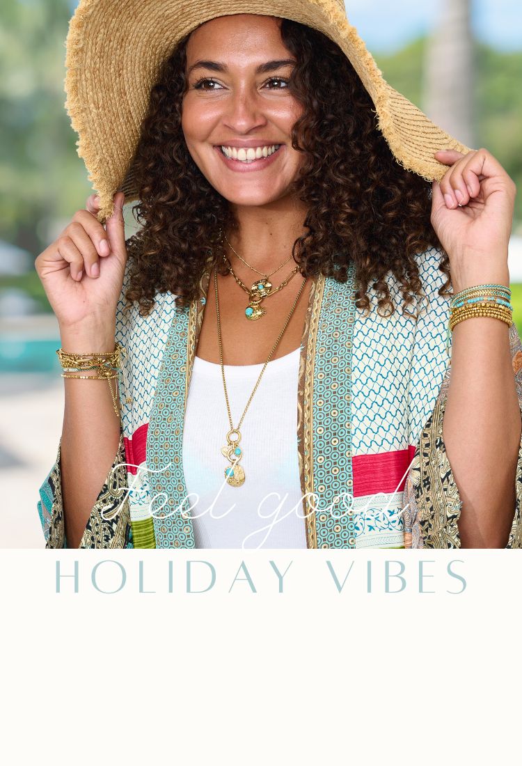 A woman stands on a sunlit beach, gazing out to sea. She wears a wide-brimmed hat and layered Boho Betty PVD jewellery—glimmering necklaces and stacked bracelets that catch the light. Her look is effortless, bohemian, and radiant—perfectly echoing the ocean breeze and carefree energy of the coast.